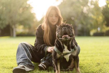 Genç bir kız çimlerin üzerinde Fransız bulldog köpeğine sarılıyor..