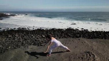 Okyanus kıyısındaki bir kayanın üstünde yoga yapan bir kız, gün batımında İzlanda 'da bir dronun manzarası.