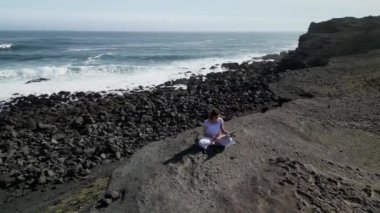 Okyanus kıyısındaki bir kayanın üstünde yoga yapan bir kız, gün batımında İzlanda 'da bir dronun manzarası.