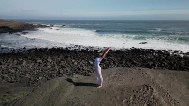 Okyanus kıyısındaki bir kayanın üstünde yoga yapan bir kız, gün batımında İzlanda 'da bir dronun manzarası.