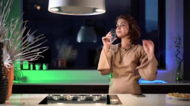 Mutfakta telefonu olan bir kadın, yeşil ışıklandırma videosunun arka planına karşı, neşeli bir elbise içinde telefon ve romantik bir sohbet bekliyor.