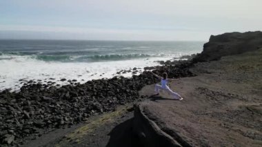 Okyanus kıyısındaki bir kayanın üstünde yoga yapan bir kız, gün batımında İzlanda 'da bir dronun manzarası.