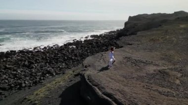 Okyanus kıyısındaki bir kayanın üstünde yoga yapan bir kız, gün batımında İzlanda 'da bir dronun manzarası.
