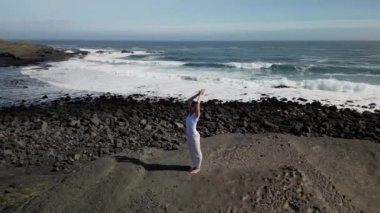 Okyanus kıyısındaki bir kayanın üstünde yoga yapan bir kız, gün batımında İzlanda 'da bir dronun manzarası.