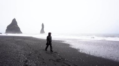 Siyah yağmurluklu bir adam İzlanda 'da siyah bir plaj boyunca yağmurda ve fırtınada yürür.