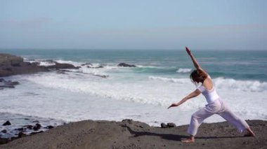 Okyanus kıyısındaki bir kayanın üstünde yoga yapan bir kız, gün batımında İzlanda 'da bir dronun manzarası.