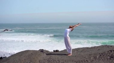 Okyanus kıyısındaki bir kayanın üstünde yoga yapan bir kız, gün batımında İzlanda 'da bir dronun manzarası.