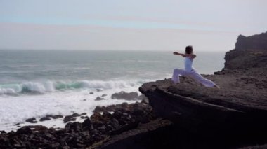 Okyanus kıyısındaki bir kayanın üstünde yoga yapan bir kız, gün batımında İzlanda 'da bir dronun manzarası.