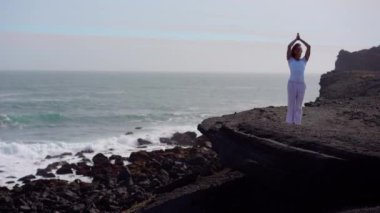 Okyanus kıyısındaki bir kayanın üstünde yoga yapan bir kız, gün batımında İzlanda 'da bir dronun manzarası.
