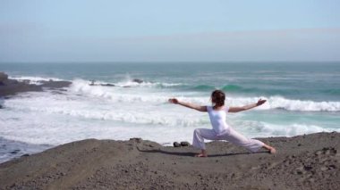 Okyanus kıyısındaki bir kayanın üstünde yoga yapan bir kız, gün batımında İzlanda 'da bir dronun manzarası.