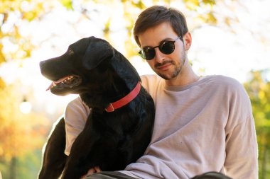 Güneş gözlüklü adam gün batımında sonbahar parkında köpeğine sarılır.