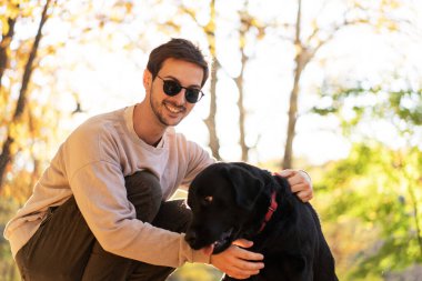 Güneş gözlüklü adam gün batımında sonbahar parkında köpeğine sarılır.