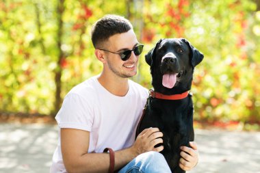 Güneş gözlüklü adam gün batımında sonbahar parkında köpeğine sarılır.