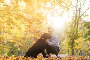 Güneş gözlüklü adam gün batımında sonbahar parkında köpeğine sarılır.