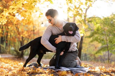 Güneş gözlüklü adam gün batımında sonbahar parkında köpeğine sarılır.