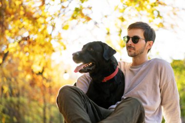 Güneş gözlüklü adam gün batımında sonbahar parkında köpeğine sarılır.