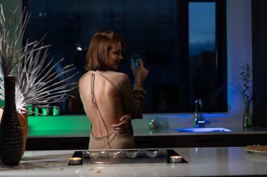 Bir kız güzel bir gece elbisesiyle mutfakta duruyor ve şampanya içiyor. Randevu bekliyor.