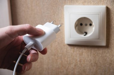 Hand turns on, turns off charger in electrical outlet on wall. White charger for phone in womans hand close-up.