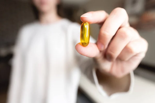 A womans hand holds an Omega-3 capsule against the background of herself in a white sweater, the background is in blur. Useful fish oil capsule tablets.