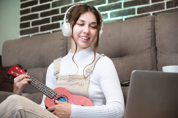 Cheerful caucasian girl playing the ukulele and looking into a laptop sits on the floor next to the sofa in the apartment. young woman learning how to play acoustic ukulele during the day at home.