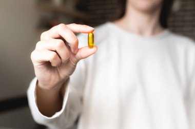 A womans hand holds an Omega-3 capsule against the background of herself in a white sweater, the background is in blur. Useful fish oil capsule tablets.