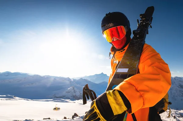 Photo of an athlete in a helmet and mask with skis on his shoulder. A young man stands in the mountains against the backdrop of the sun and looks at the camera while holding skis.