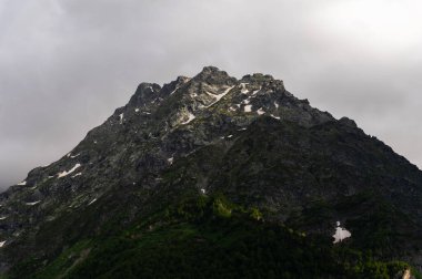 Rocky peak of a mountain with snow, sunlight shines on the crest of the peak, there is a forest at the base of the mountain. Full frame close-up.