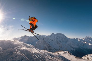 Kış tatili kayak merkezinde. Kayakçı hızla havada uçar ve arka plandaki kar örtülü dağlarda helikopter kayağı yapar..