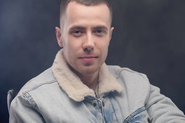 portrait of a young man in a denim jacket, looking straight at the camera, haze around, studio shot.