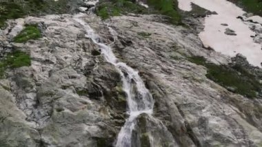 Buzuldan kayaların üzerinden akan dağ şelaleleri ile hava manzarası yakın uçuştadır. Yeşil bir dağ geçidine düşen su. Üç eksende hareket.