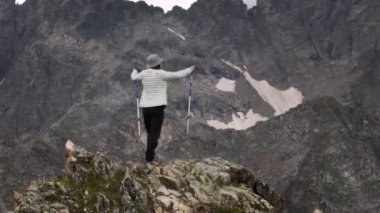 Genç bir kadın yolcunun telefoto görüntüsü, kar örtülü destansı dağların zemininde bir uçurumun tepesinde duruyor. Zirveye ulaşan zafere uzanıyor..