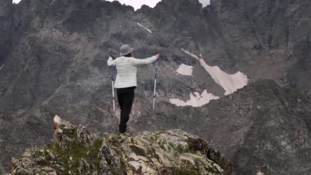 一个年轻女性旅行者的空中心灵感应画面站在悬崖顶上 背靠着白雪覆盖的史诗山脉 高举胜利的双手到达山顶 — 图库视频影像