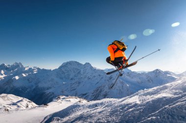 Kayakçı mavi gökyüzünün ve kar kaplı dağların arka planında atlar. Serbest stil kayakçı tam dönüşle aynı anda çapraz kayak yapan bir helikopter kullanır..