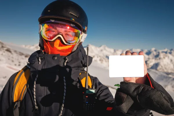 背景には山のある空のリフト券を持っているスノーボーダー 冬の装備をカメラを見て若いスキーヤーの手の中に空白のスキーパス スキーの入場料を示す概念 — ストック写真