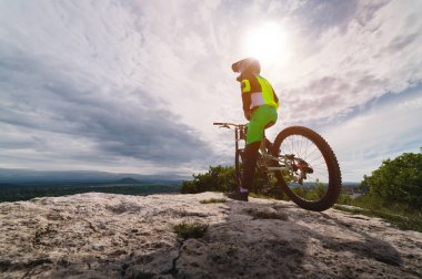 Spor giyimli genç bir adam bir dağ bisikletinin yanında duruyor ve bir yarışma için güzergah seçerken yan tarafa bakıyor. Sporcu antrenmandan sonra dinleniyor..