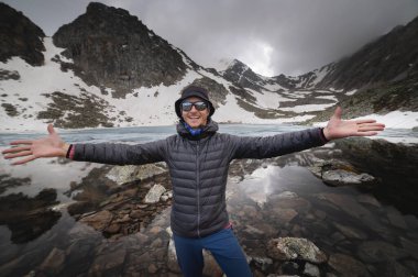 Kolları açık genç ve güçlü bir adam kameraya bakıyor. İnsanlar dağlarda ve doğada kendilerini iyi, mutlu ve özgür hissediyorlar. Dağ gölünde yürüyüş yapan mutlu adam.