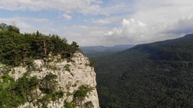 Uzun saçlı beyaz bir adamın havadan görünüşü, yeşil ormanlarla kaplı bir vadideki bir vadinin arka planına karşı yüksek bir kayanın tepesindeki uçurumun kenarında duruyor. Seyahat videosu.