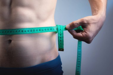 Gri bir arka planda, belinde bir mezura tutan genç bir sporcunun gövdesi. Genç bir adam belini mezurayla ölçer, yakın plan..