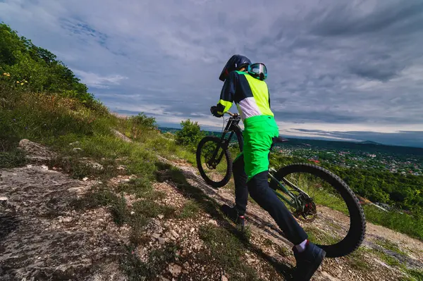 A mountain biker carries a bicycle in a forested landscape of summer mountains. MTB track for cycling. Outdoor sports, the guy carries the bike to the top