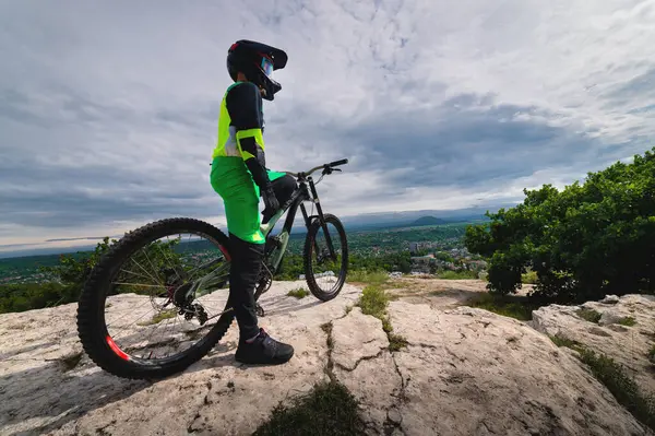 A wide angle of a young mountain bike rider in a helmet and a protective suit on his mountain bike stands on a rock against the backdrop of clouds and a panorama of the private sector of the city in