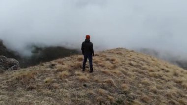 Aşağı ceketli ve şapkalı beyaz bir adam uçurumun kenarında dikiliyordu. Dağlarda, bulutların altında..