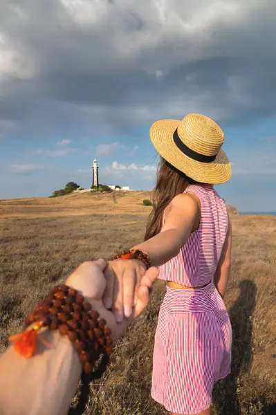 A young woman is traveling through the countryside with her man when they stumble upon a field with a lighthouse. Free girl and guy walk hand in hand at sunset, holding hands, unrecognizable people.