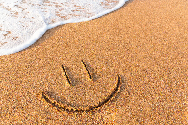 A smile painted on the sand next to a foamy wave. Sandy beach with a friendly message written on it