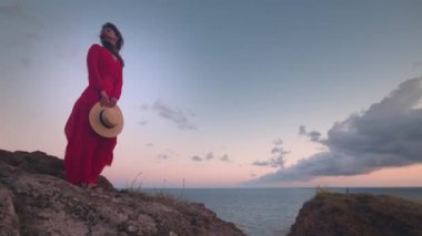Gün batımından sonra rüzgarlı bir havada elinde şapkasıyla kayalık bir sahilde duran uzun kırmızı elbiseli çekici beyaz bir kadın. Deniz kıyısındaki kız