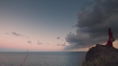 Yukarı sahilde gizemli romantik bir kadın duruyor ve uzaklığa bakıyor. Bulutlu bir havada güneşin doğuşunun ya da batışının görüntüleri.