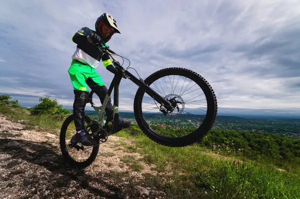 stock image an athlete performs a stunt while standing on a wheelie on a rock, overlooking green forests and mountains. cycling extreme sport, man in full protection of the whole body and head