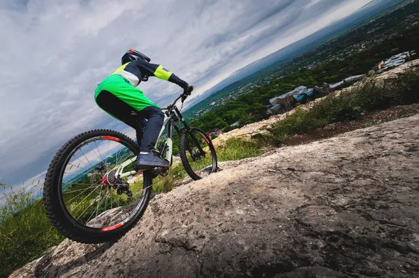 A young cyclist rides off a cliff on his mountain bike against the backdrop of the private sector of the village. Wide angle low angle with obstructed horizon.