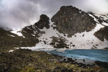 Yüksek bir dağ manzarası, karlı bir dağ sırası ve önü buzla kaplı bir göl. Bulutlu havada taş tepeler.