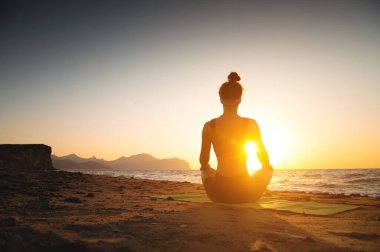 A woman meditates on the coast by the sea, rear view. Place for text. Yoga, health harmony, silhouette of a young woman on the beach at sunset or dawn. clipart