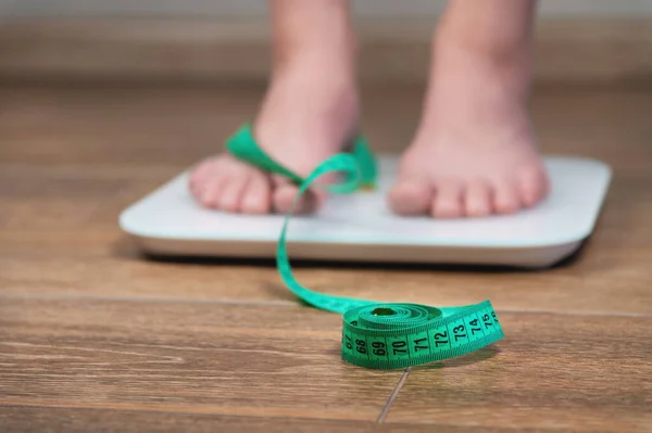 A girl with swollen feet uses a scale next to a measuring tape on a wooden floor. Unrecognizable woman weighing herself.